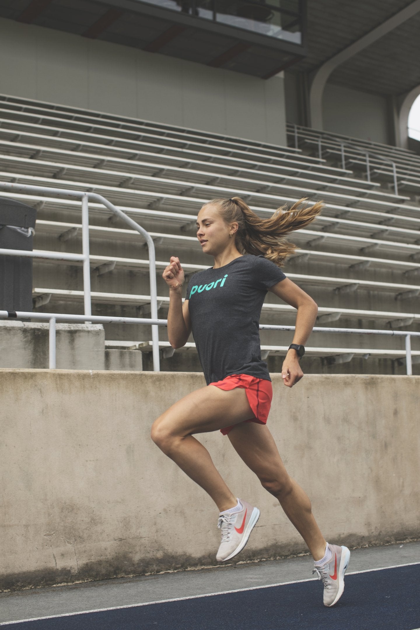 Female runner sprinting on an outdoor athletics track wearing a dark Puori-branded t-shirt and coral shorts, empty stadium bleachers in the background.